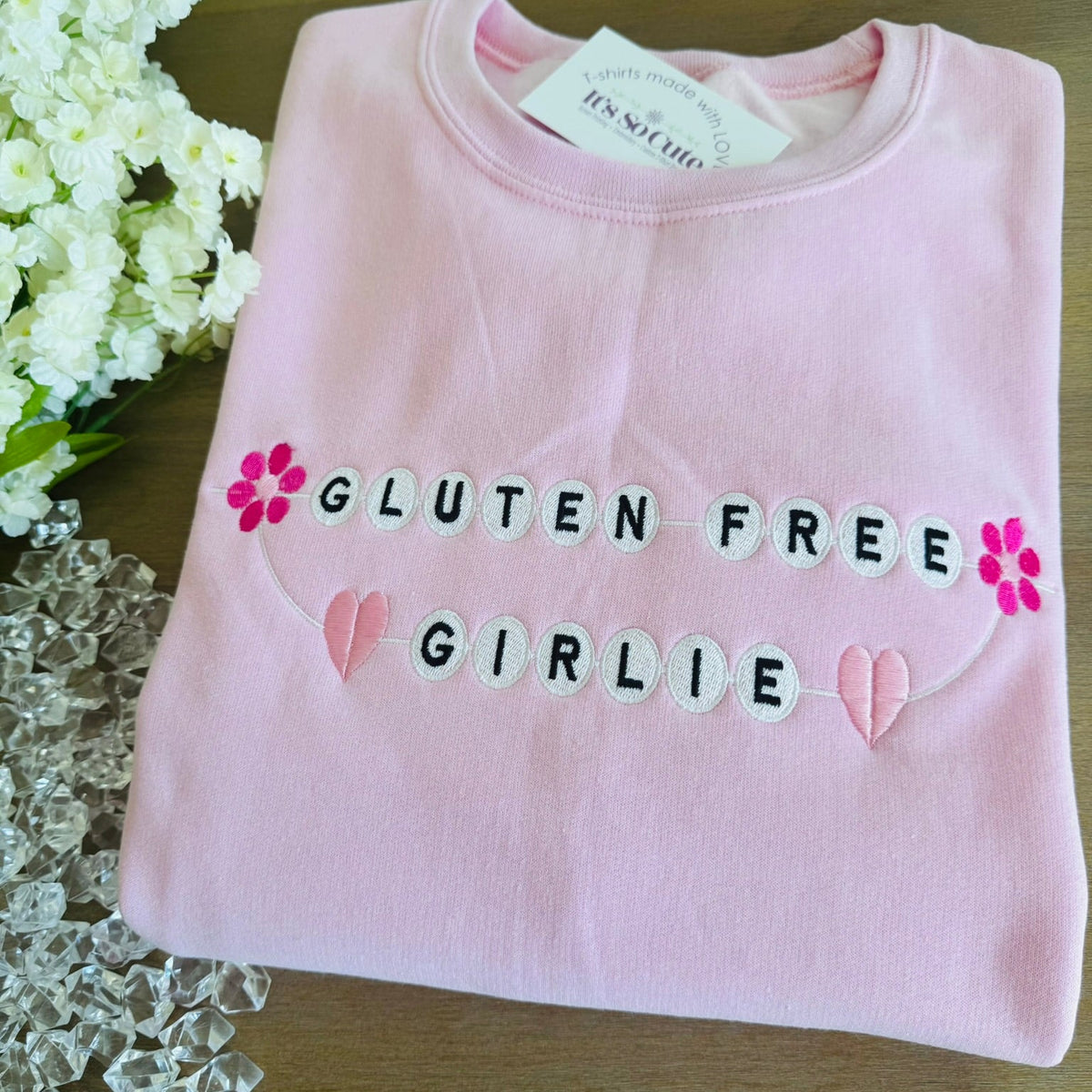 A folded light pink sweatshirt on a wooden surface, featuring the words 'GLUTEN FREE GIRLIE' in black embroidered lettering with white letter tiles, pink flower accents, and pink heart details.