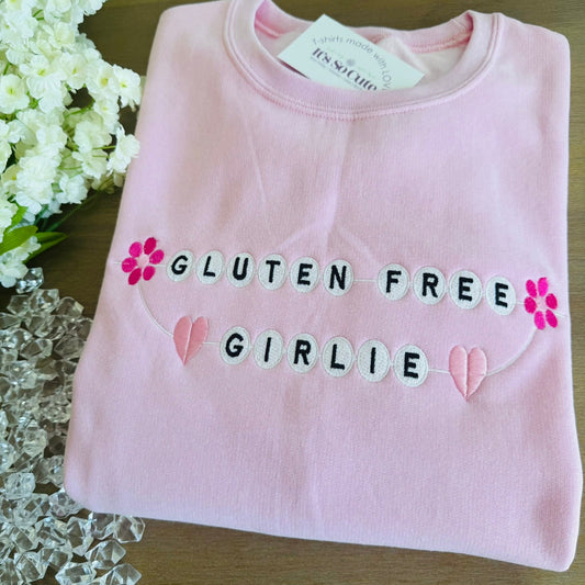 A folded light pink sweatshirt on a wooden surface, featuring the words 'GLUTEN FREE GIRLIE' in black embroidered lettering with white letter tiles, pink flower accents, and pink heart details.