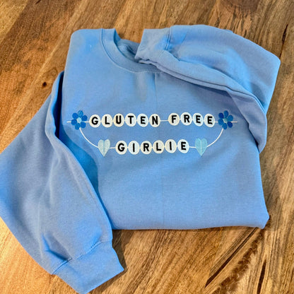 A light blue crewneck sweatshirt with a 'GLUTEN FREE GIRLIE' graphic embroidered on the front, laid flat on a wooden surface.