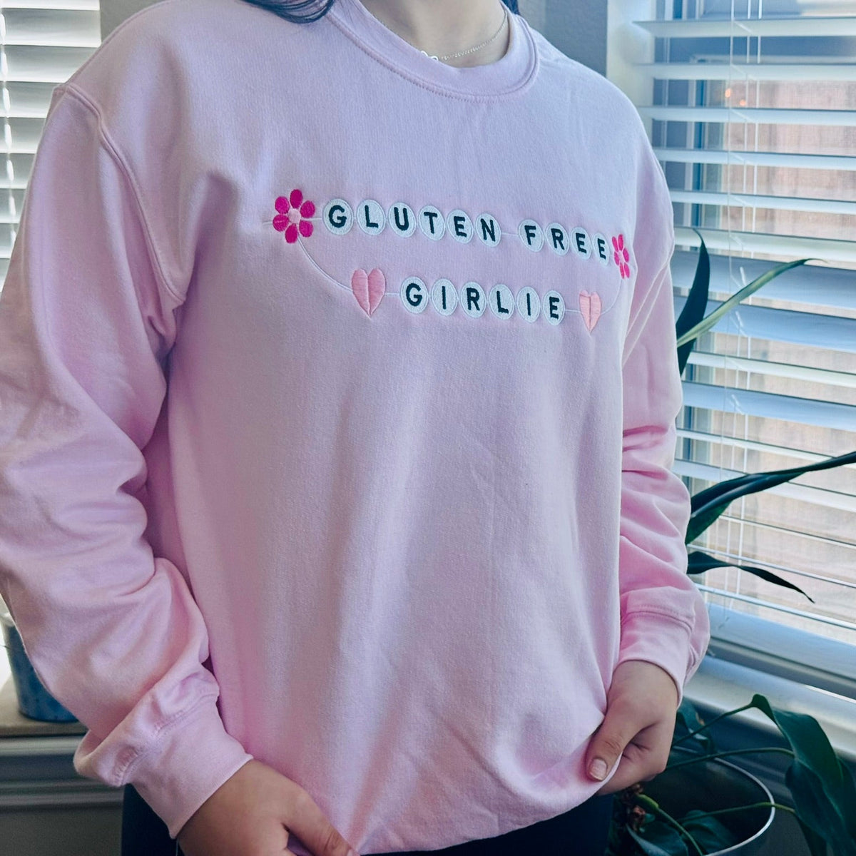 A person wearing a light pink long-sleeve sweatshirt with 'GLUTEN FREE GIRLIE' text and small pink flower and heart embroidery on the chest, standing indoors near a window with blinds.