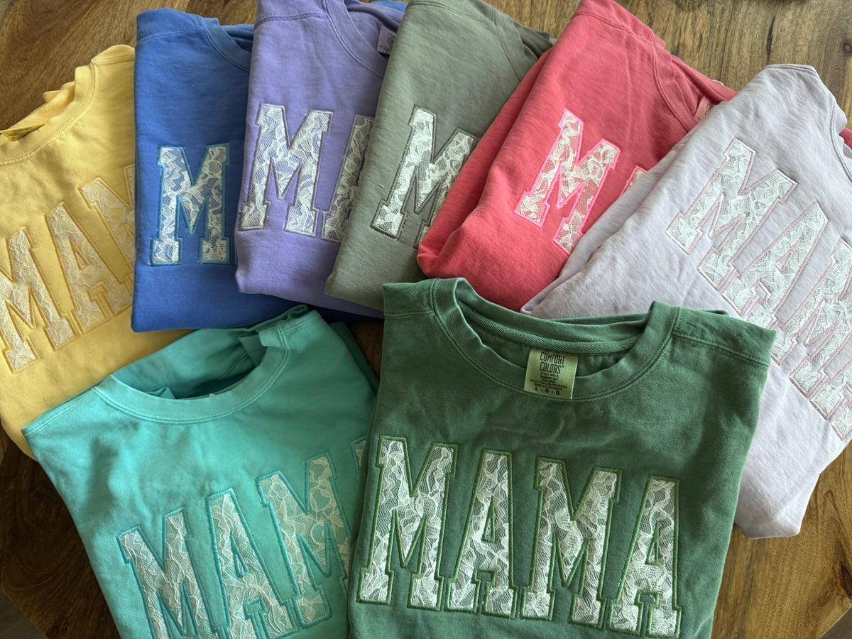 A group of folded sweatshirts arranged on a wooden surface, each featuring a large distressed 'MAMA' applique across the front in a contrasting color. The shirts shown include yellow, blue, lavender, coral pink, white, and green.
