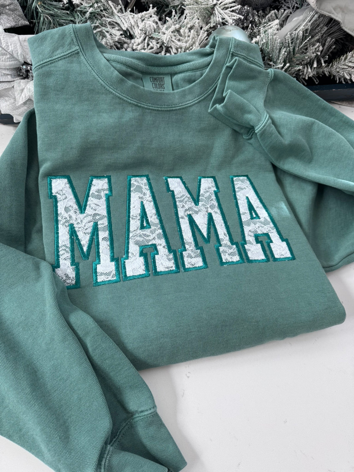A folded sage green sweatshirt featuring the word 'MAMA' in large white varsity-style letters with a teal outline, styled on a white surface with a silver tinsel decoration in the background.
