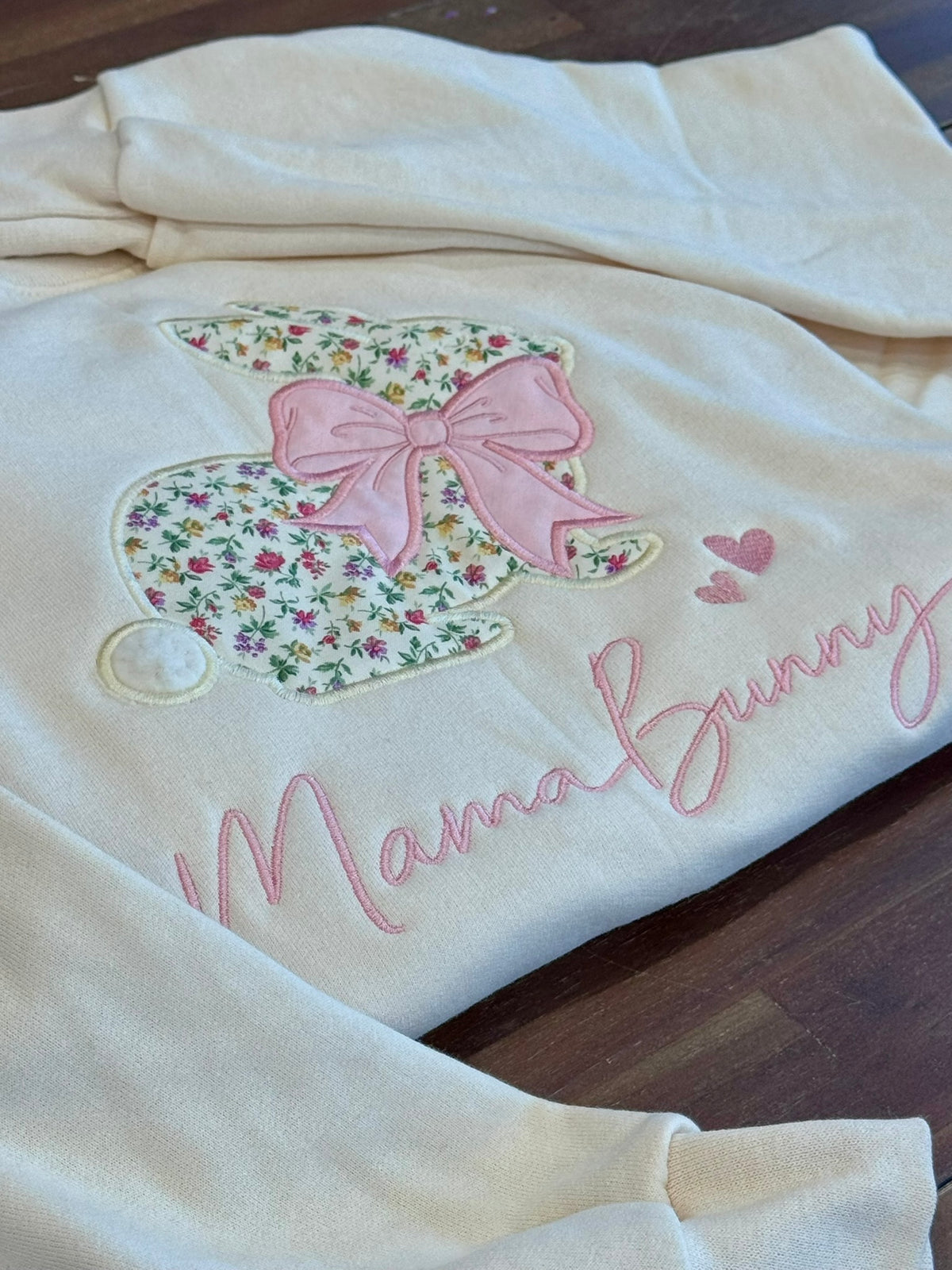 An off-white fleece sweatshirt featuring an applique bunny design with a pink bow and floral pattern, alongside the embroidered text 'Mama Bunny' in pink script, laid flat on a dark wood surface.
