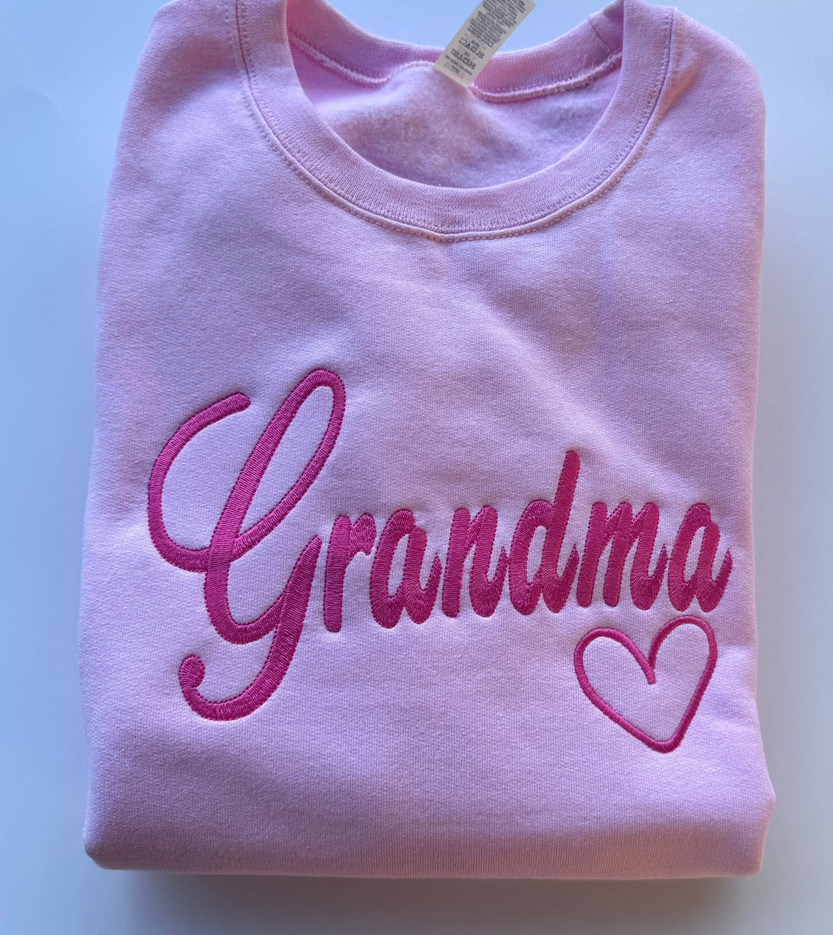 A folded light pink crewneck sweatshirt featuring the word 'Grandma' and a heart embroidered in bright pink on the front.