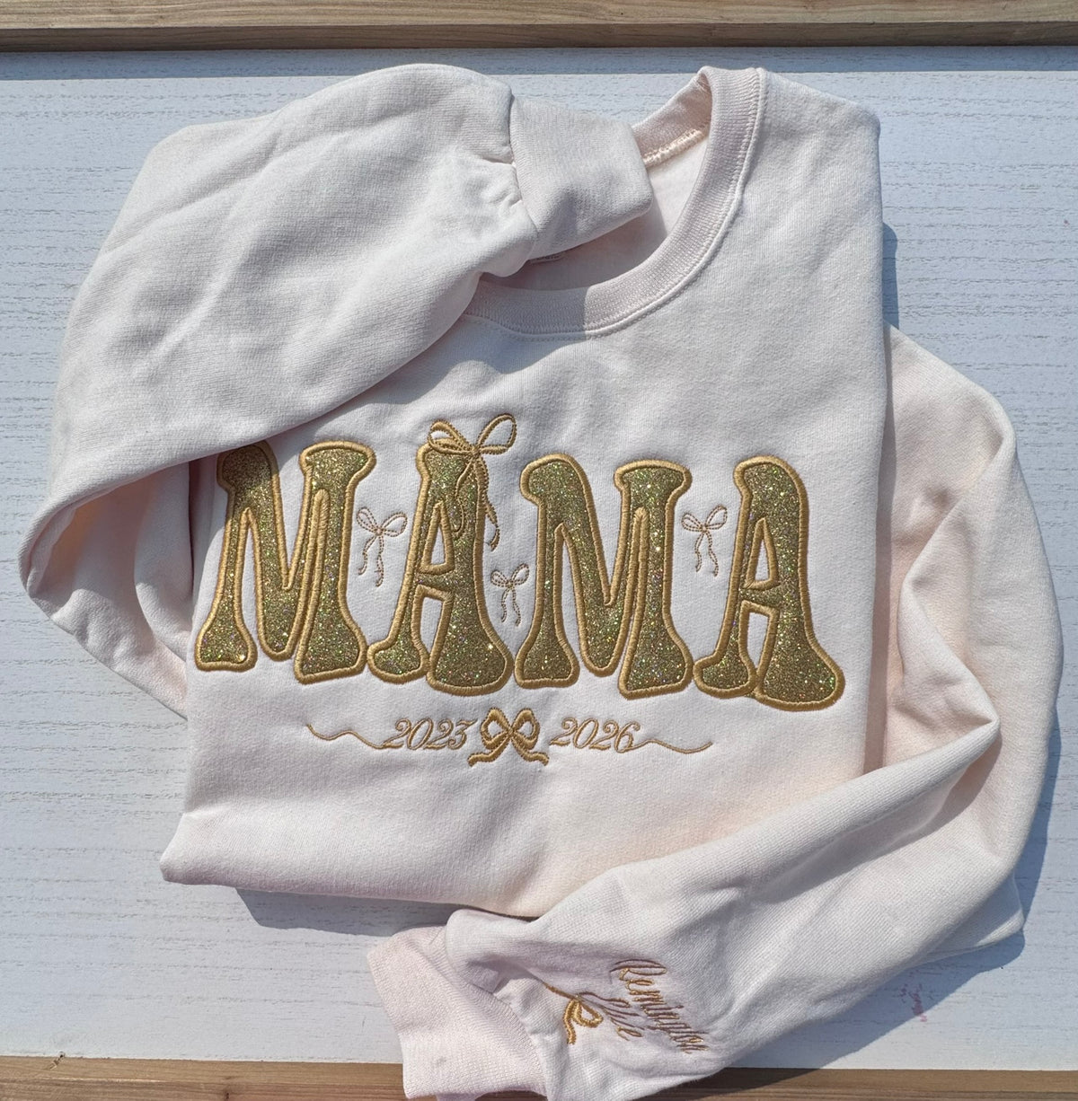 A light pink long-sleeve sweatshirt with 'MAMA' embroidered in glittery gold applique lettering, laid flat on a white wooden surface.