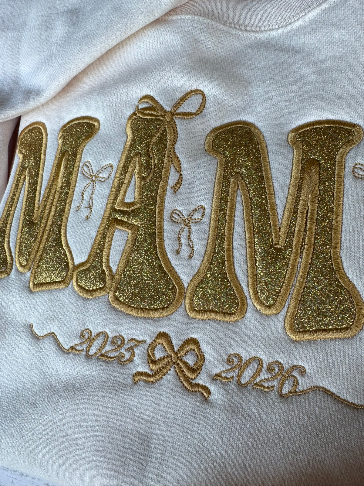A close-up of a white sweatshirt featuring a glittery gold applique 'MAMA' design with small bow details and the years '2023' and '2026' embroidered below.