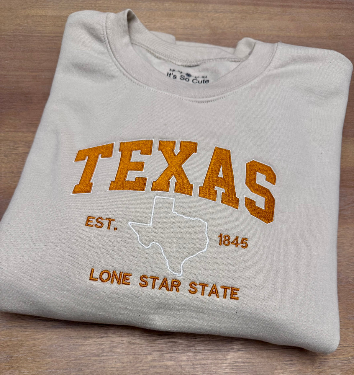 A folded light beige crewneck sweatshirt with 'TEXAS' and 'LONE STAR STATE' embroidered in orange, featuring a white outline of the state of Texas in the center, laid flat on a wooden surface.