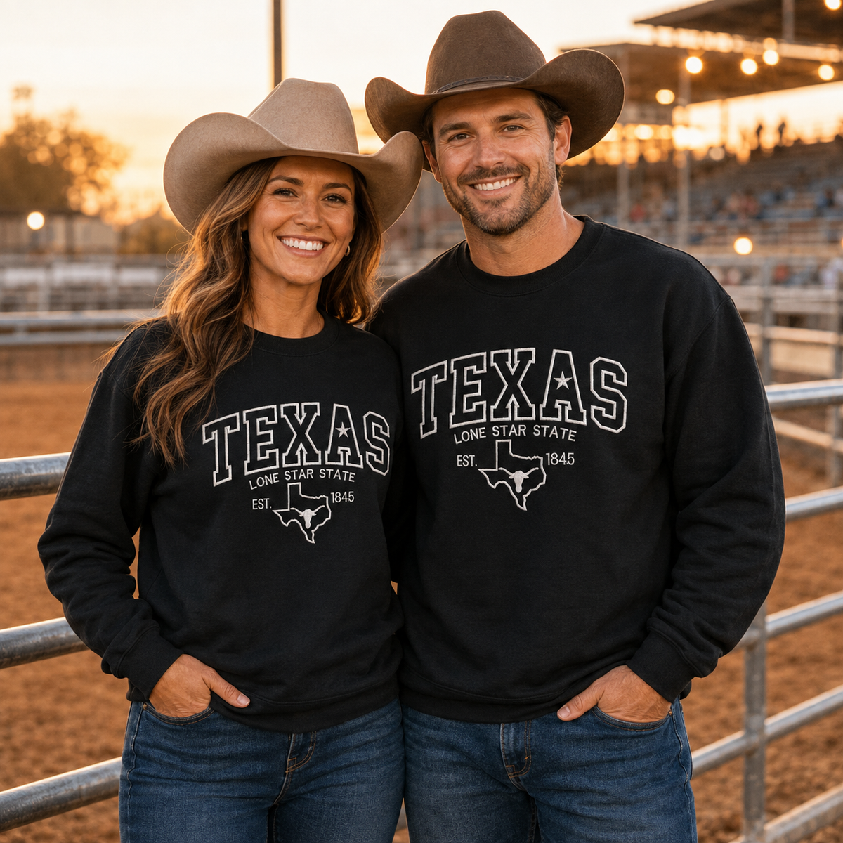 A man and a woman standing together in a rodeo arena at sunset, both wearing matching black crewneck sweatshirts with white 'TEXAS LONE STAR STATE EST. 1845' embroidered and cowboy hats.