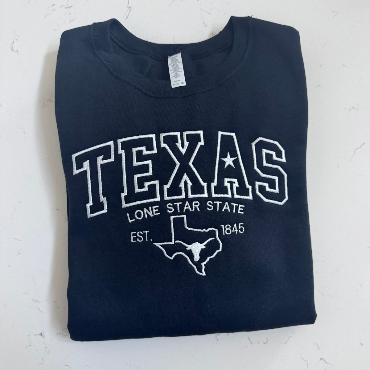 A folded black t-shirt with a white embroidery that reads 'TEXAS LONE STAR STATE EST. 1845' and a Texas state outline, laid flat against a white textured background.