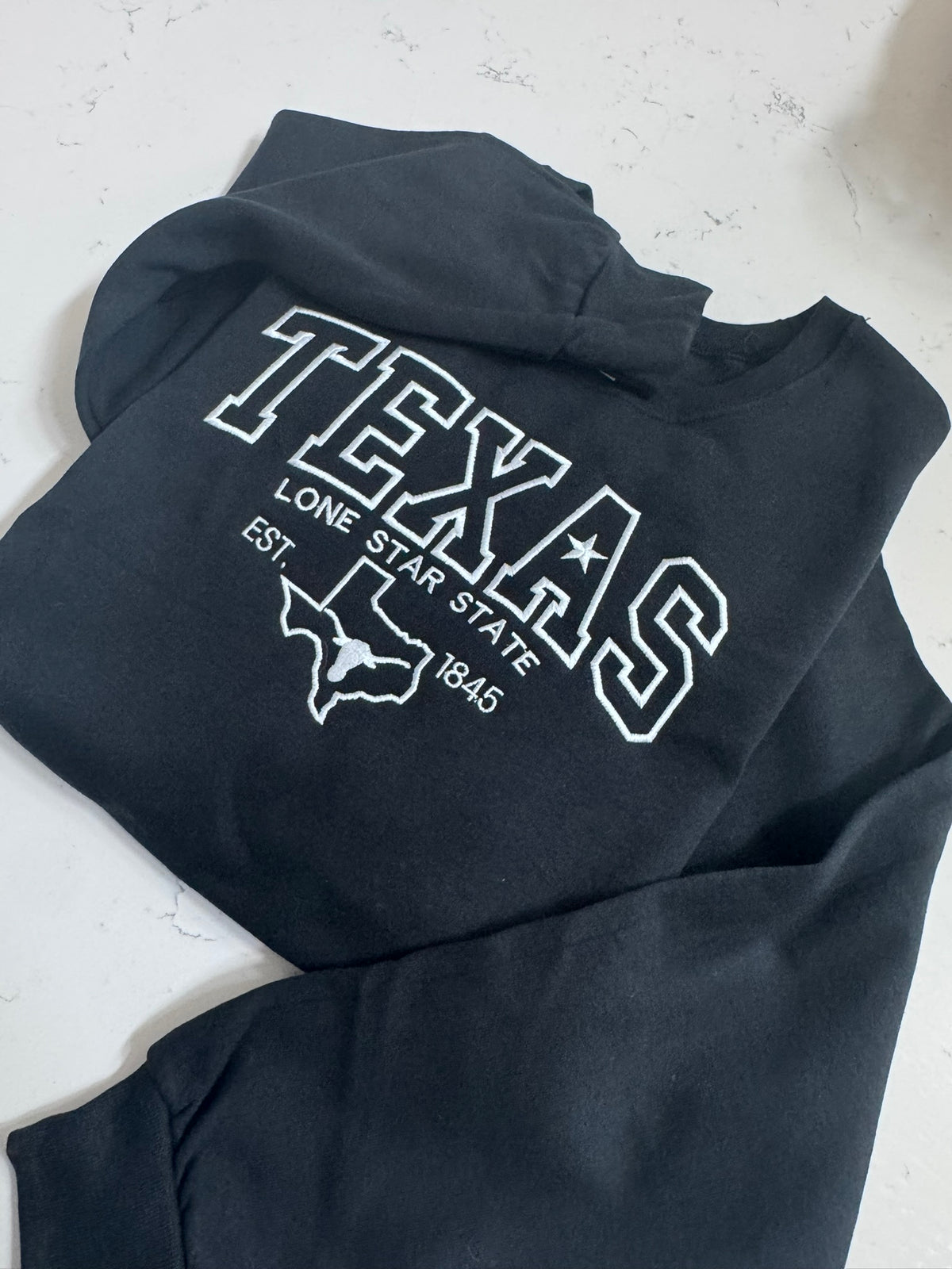 A folded black sweatshirt with a white embroidery reading 'TEXAS LONE STAR STATE EST. 1845' and a Texas state outline, laid flat on a light surface.