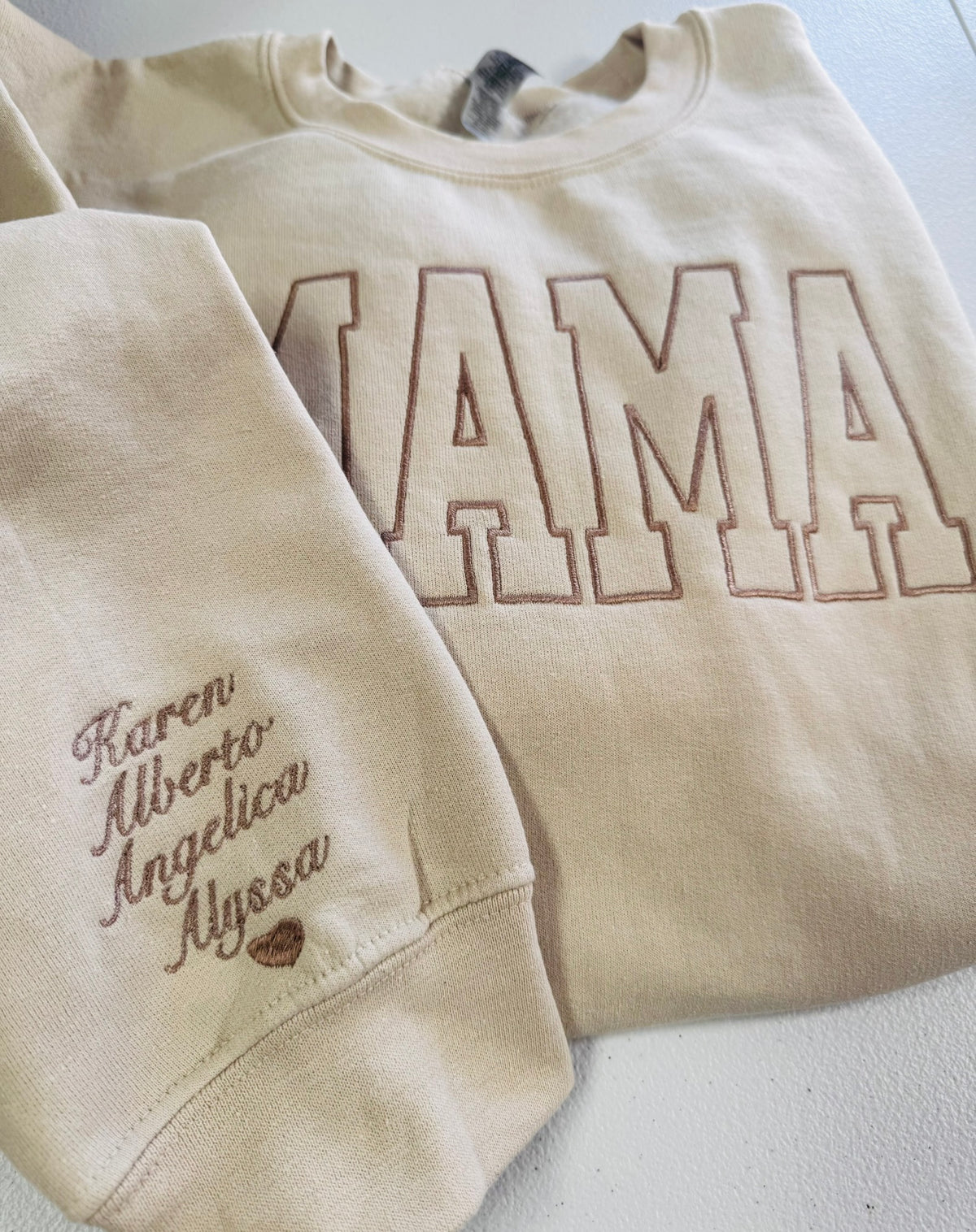 A beige crewneck sweatshirt with 'MAMA' outlined in brown embroidery across the chest and custom names embroidered in brown script on the left sleeve.