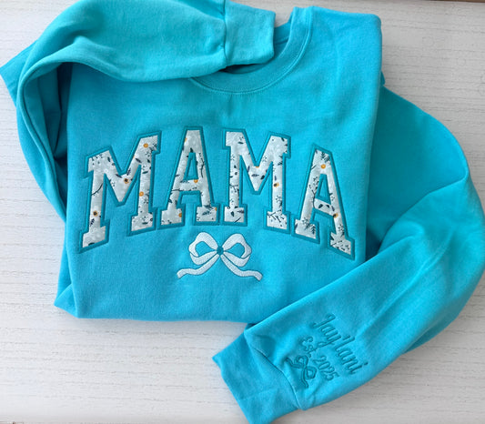 A turquoise long-sleeve sweatshirt laid flat on a white wood background. The front has large arched 'MAMA' applique with a floral pattern and a white bow embroidery below.