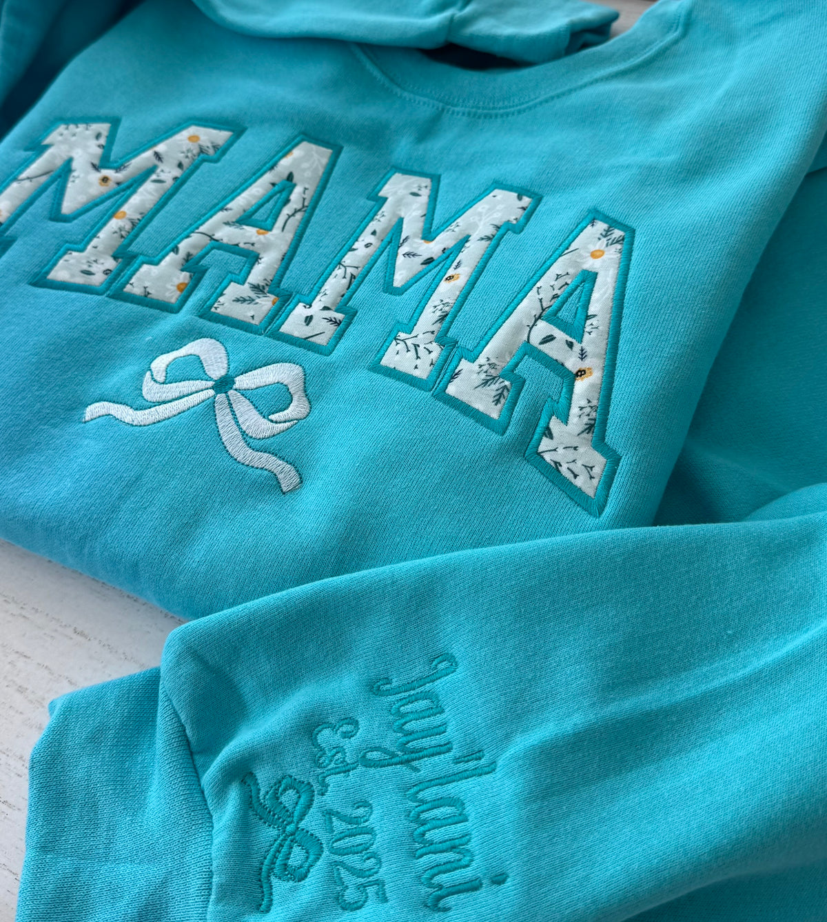 A turquoise crewneck sweatshirt featuring 'MAMA' in large white applique letters with a floral pattern and a white bow embroidery on the chest, with embroidered in teal script on the sleeve.